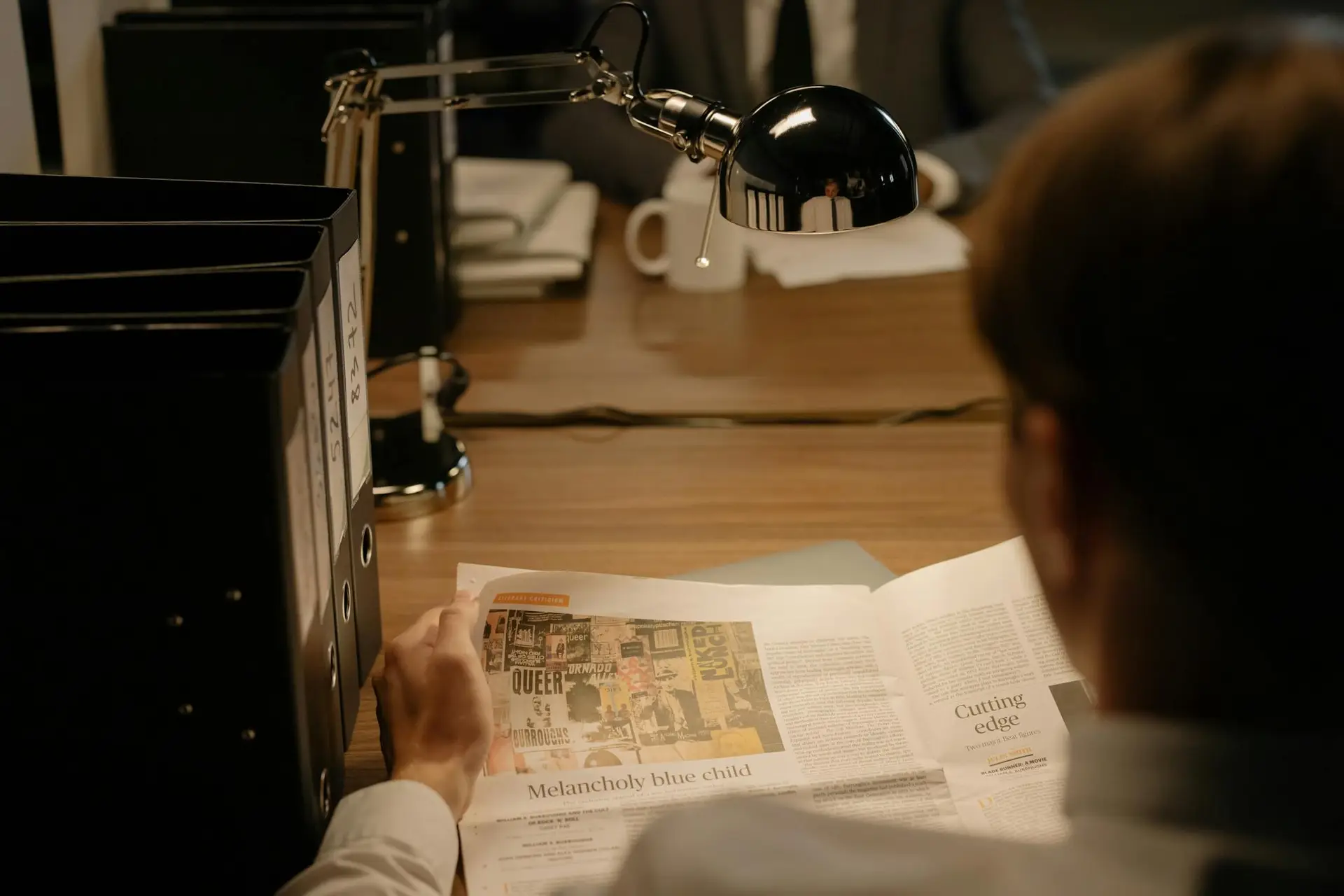 A person reads a newspaper at a desk with a lamp, suggesting investigation work.